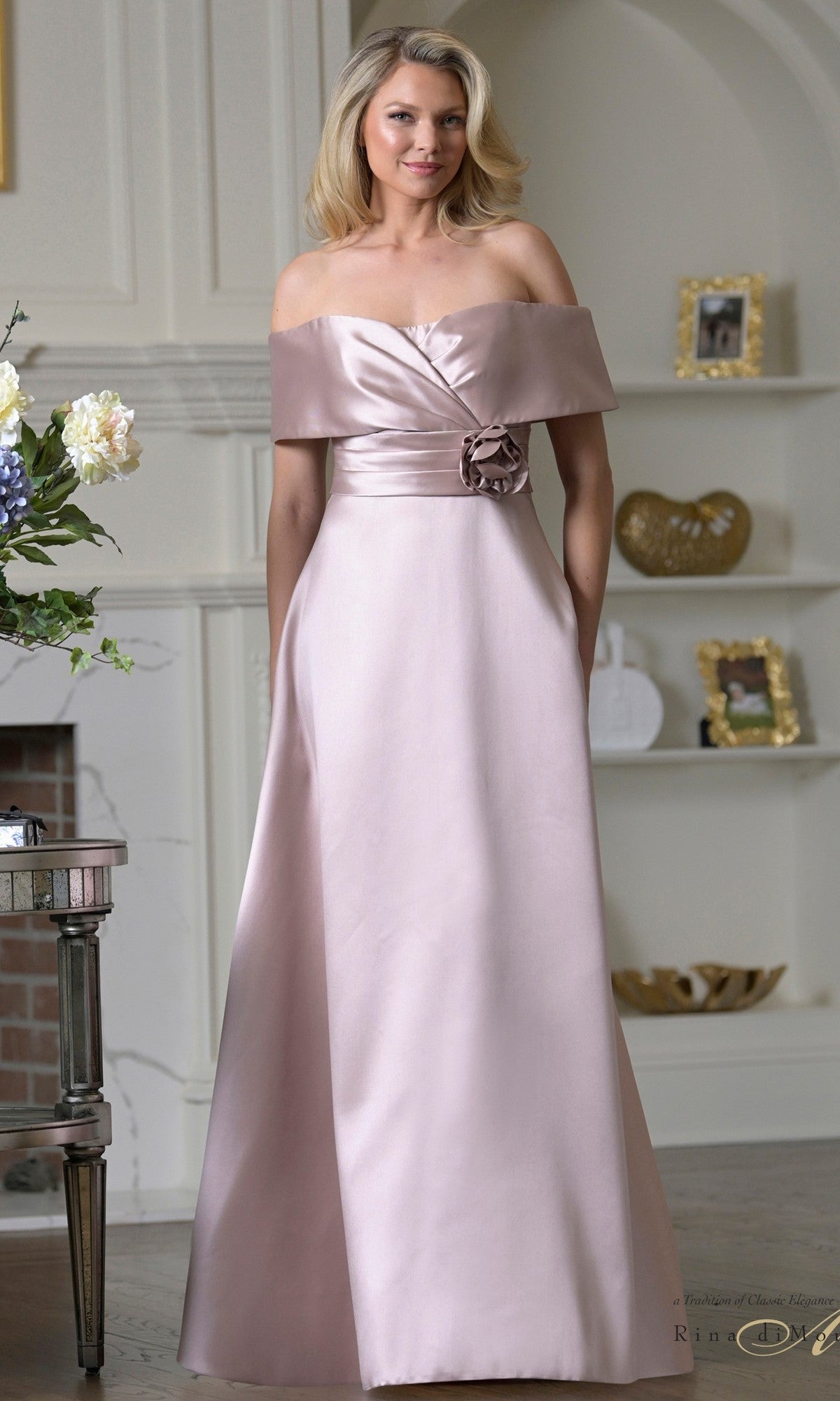A woman stands in a decorated room wearing the Rina Di Montella RD3429 long formal dress, featuring an off-the-shoulder design, light pink color, and a floral detail at the waist.